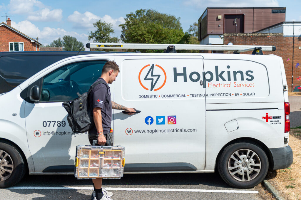 Hopkins Electricals electrician carrying a parts case to a white van with the Hopkins logo, website and NICEIC badge on the side