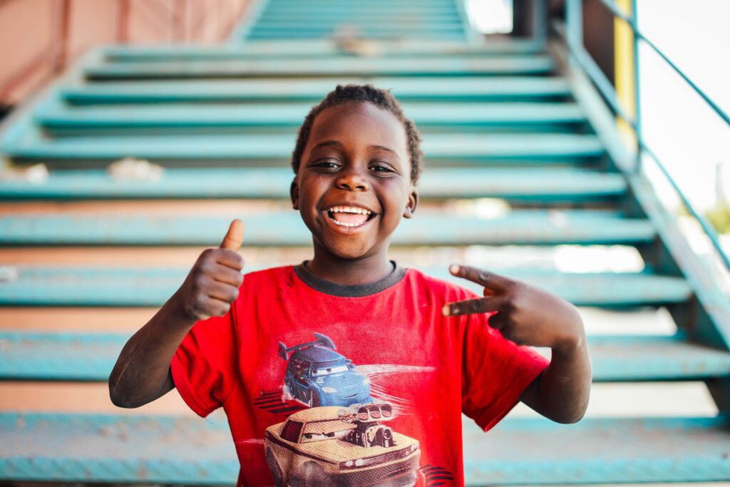 child with a red t-shirt grinning with their right hand showing thumbs up